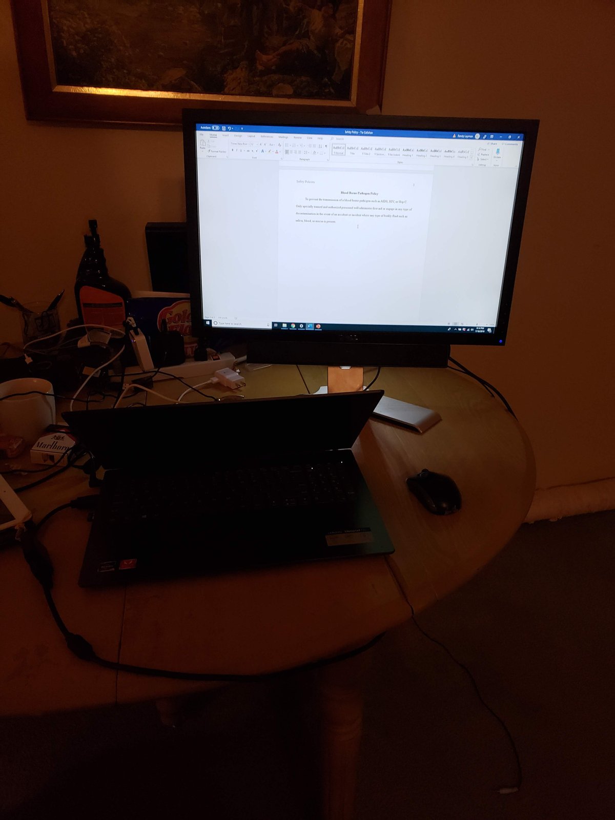 Desk with a monitor showing a Word document, a laptop and a mouse on a round table, with a framed painting on the wall above.