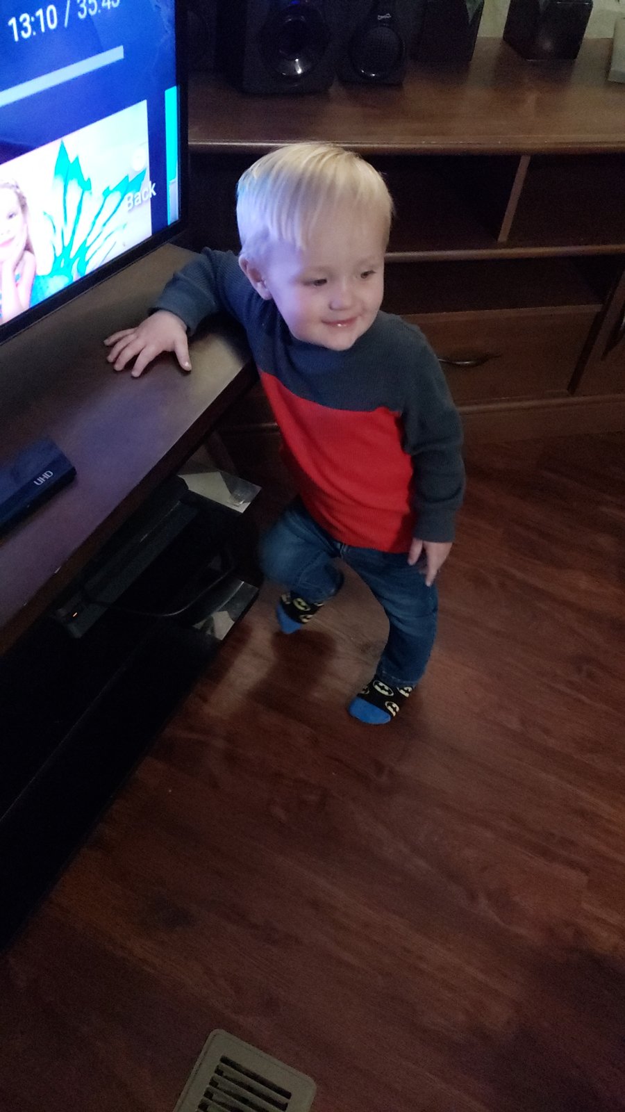 Smiling toddler with blond hair in a red and gray shirt standing beside a TV stand in a living room.