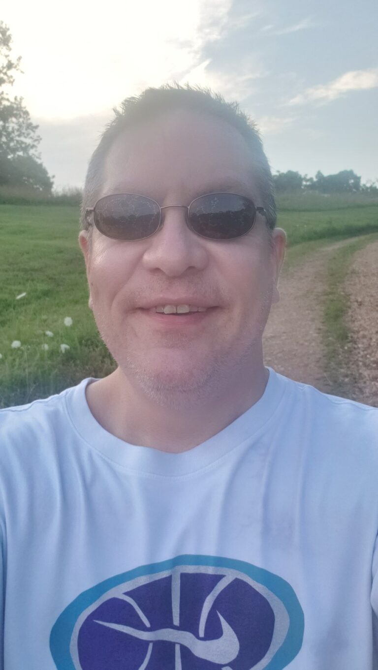Selfie of a smiling man in sunglasses and a white T-shirt outdoors, with a grassy field and dirt road behind him.