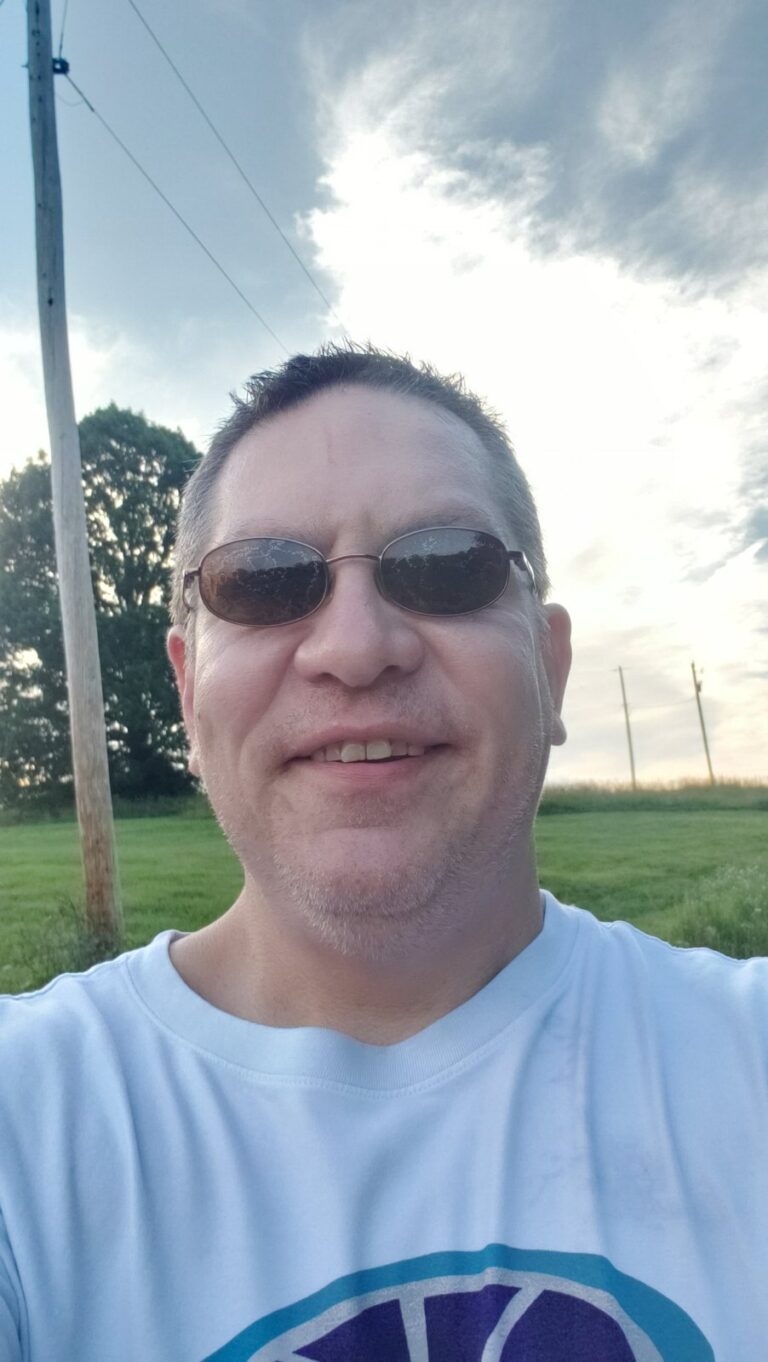 Selfie of a smiling man in sunglasses and a white T-shirt outdoors, with a utility pole, trees and a cloudy sky behind him.